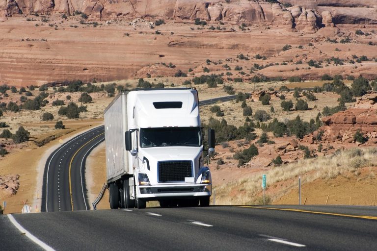 A white semi-truck drives on a curved highway through a desert landscape with rocky hills and sparse vegetation under a clear sky.