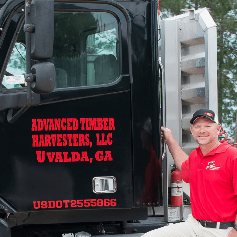 Man in red shirt and black cap stands beside a black truck labeled "Advanced Timber Harvesters, LLC Uvalda, GA." The truck door is open, and a fire extinguisher is visible.