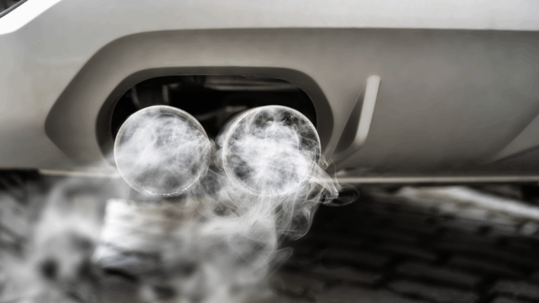 Close-up of a vehicle’s dual exhaust pipes emitting white smoke, with cobblestones visible in the background, illustrating the hidden costs of engine idling.
