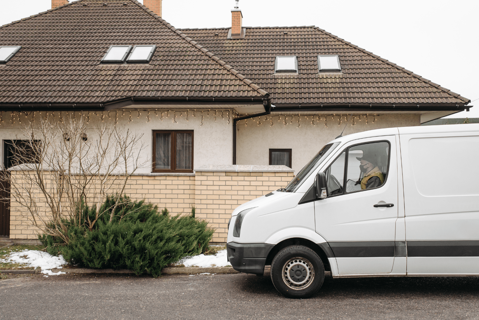 A white delivery van is parked in front of a beige house with a tiled roof and Christmas lights. Some snow and greenery are visible near the driveway, reminding us that the costs of engine idling—even during quick stops—can add up over time.