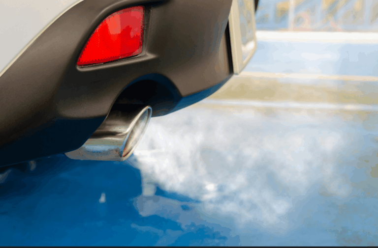 Primer plano del tubo de escape de un coche que emite humos visibles sobre una superficie azul, con la luz trasera y el parachoques parcialmente visibles, lo que ilustra los costes del motor al ralentí, que a menudo se pasan por alto.