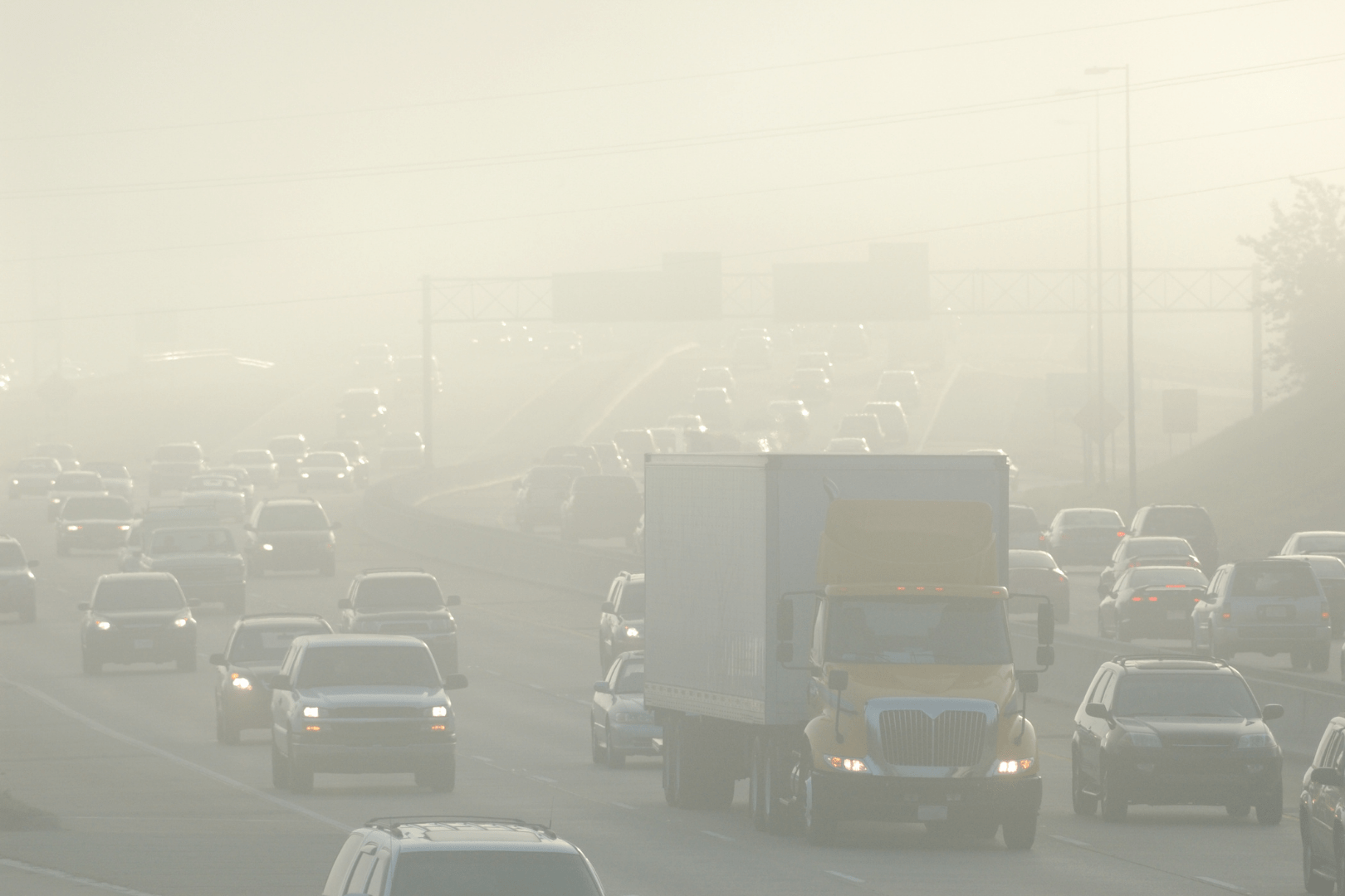 Dense fog covers a busy multi-lane highway filled with cars and trucks, reducing visibility and creating hazardous driving conditions. As vehicles slow down or idle in traffic, the hidden costs of engine idling add to the challenges faced by drivers.