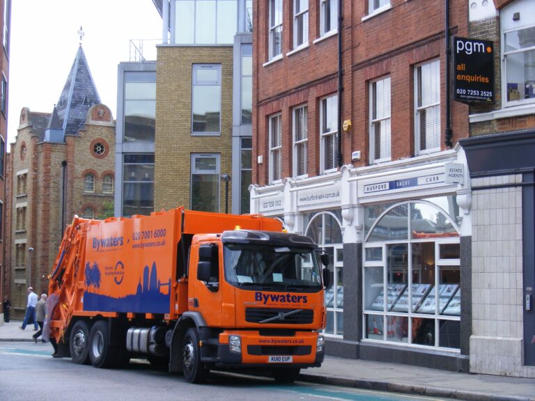 Orange Bywaters refuse truck parked on a city street near brick buildings and a design agency.