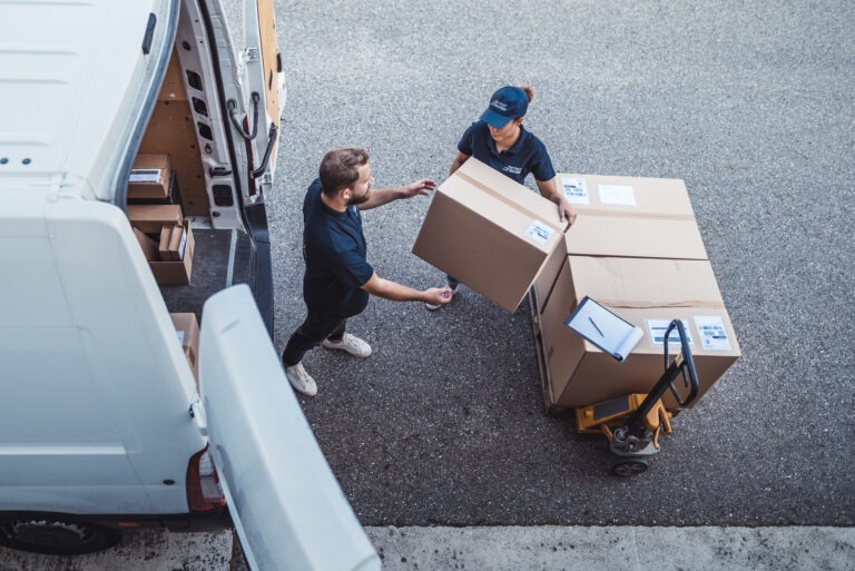 Two people in uniforms unload large cardboard boxes from a delivery van onto a hand truck.