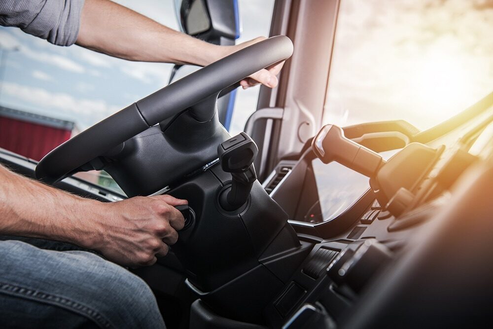 A person is starting a vehicle, hand turning the ignition key as sunlight streams through the window. The steering wheel and dashboard, sleek with modern facilities, are clearly visible.