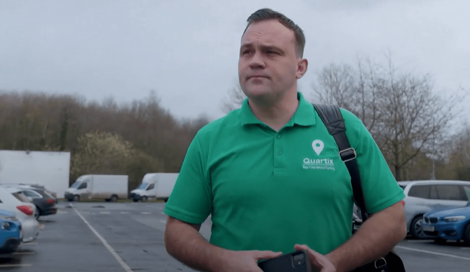 Person in a green polo shirt holding a phone and bag walks through a parking lot with cars and vans, surrounded by trees under a cloudy sky.