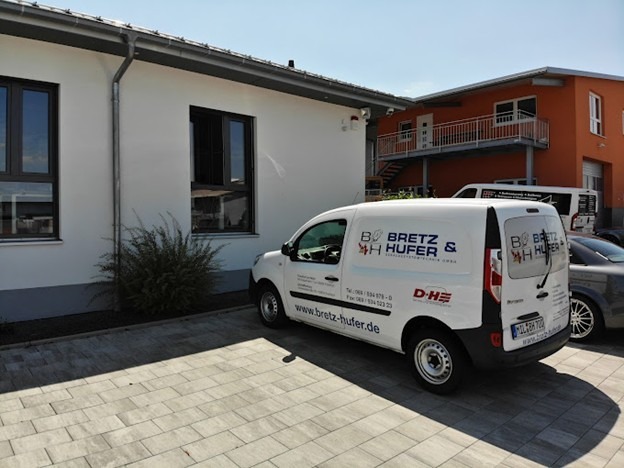 A white van with "Bretz & Hufer" branding is parked on a paved driveway outside a white building with large windows.