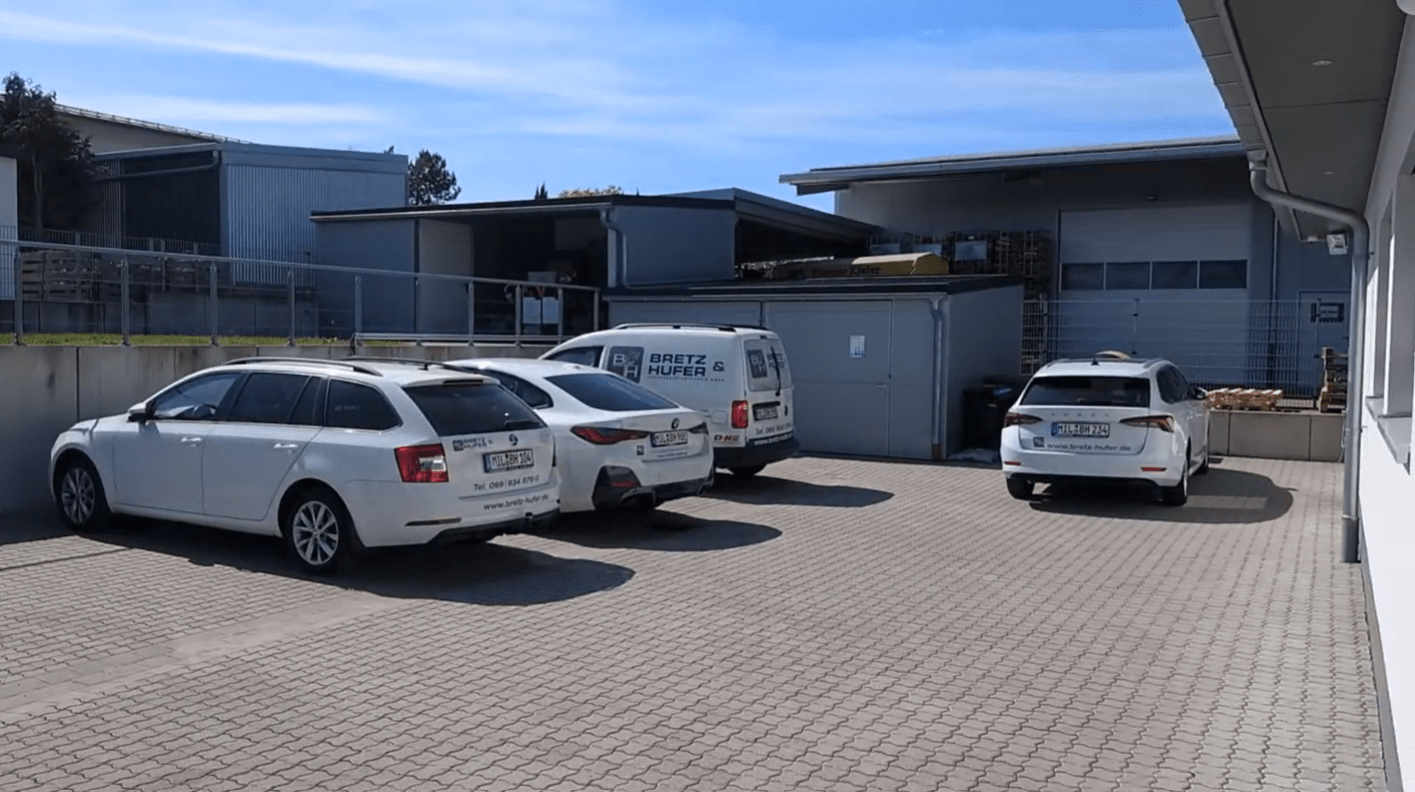 Three white vehicles, including two station wagons and a van labeled "BRETZ & HUFER," are parked on a paved lot beside industrial buildings under a clear sky.