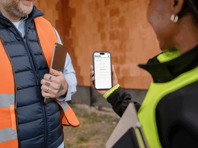 Two people in safety vests stand outside; one shows a smartphone with a checklist app to the other, who is holding a tablet.