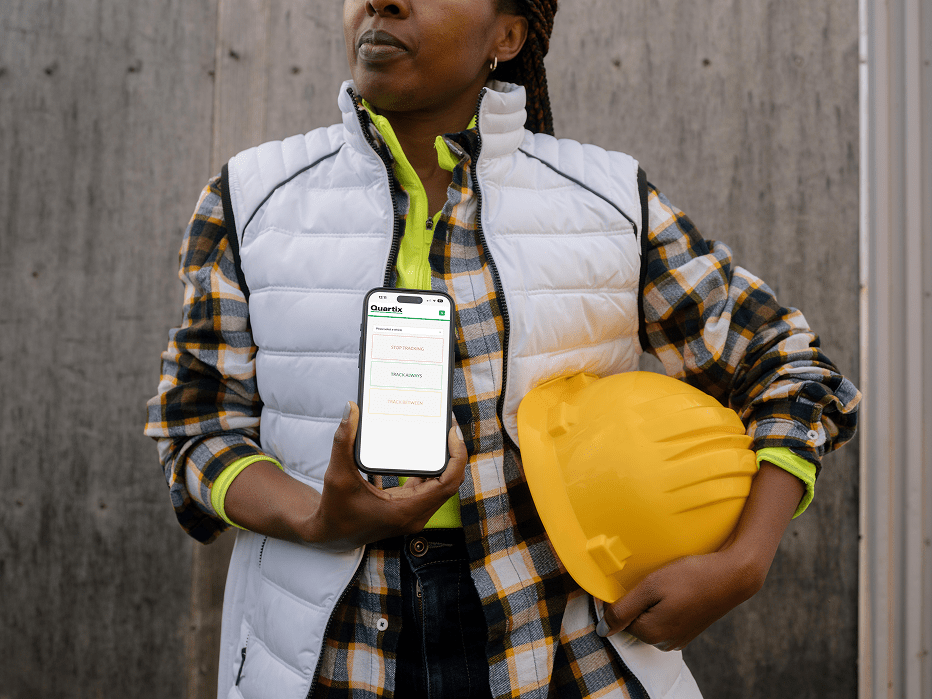 A person in a plaid shirt and vest holds a yellow hard hat and displays a smartphone with the Quext app open, standing in front of a wooden wall.