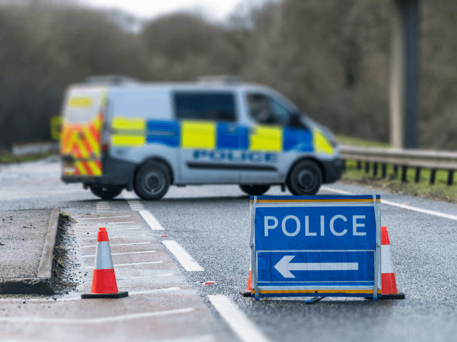 A blurred police van blocks a road, with traffic cones and a blue sign reading "POLICE" and an arrow directing traffic to the left.