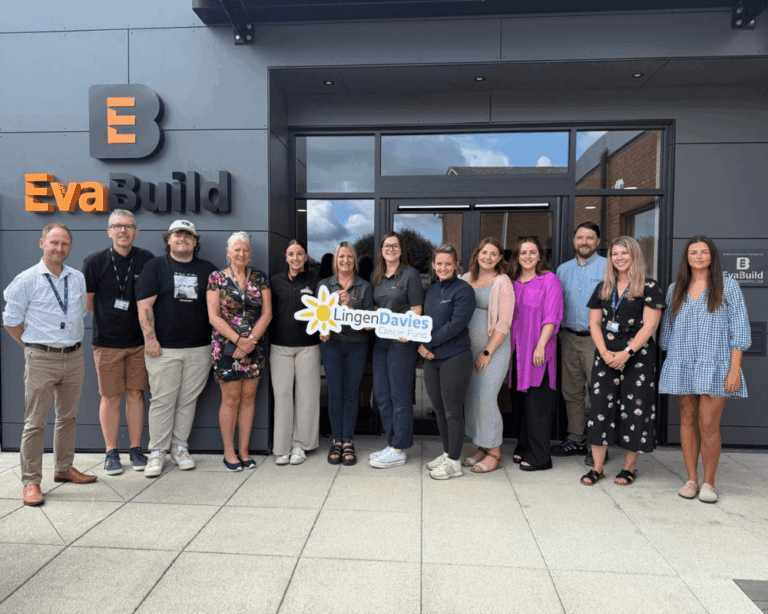 A group of thirteen people stand outside the EvaBuild building, holding a sign for Lingen Davies Cancer Fund and posing for a group photo.