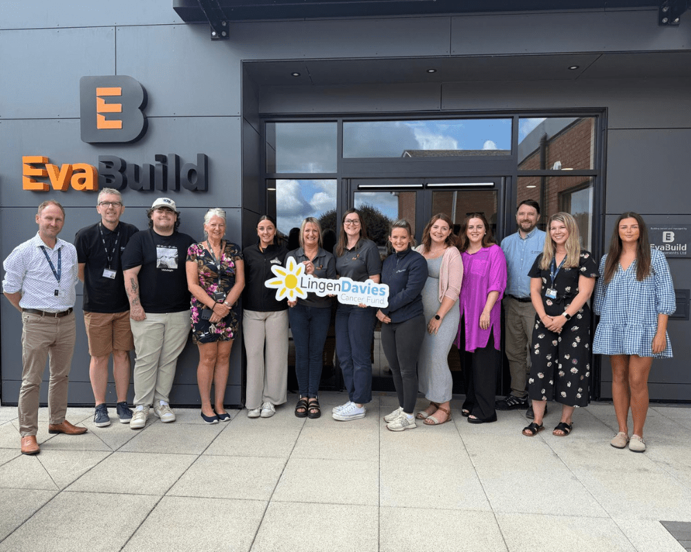 A group of thirteen people stand outside the EvaBuild building, holding a sign for Lingen Davies Cancer Fund and posing for a group photo.