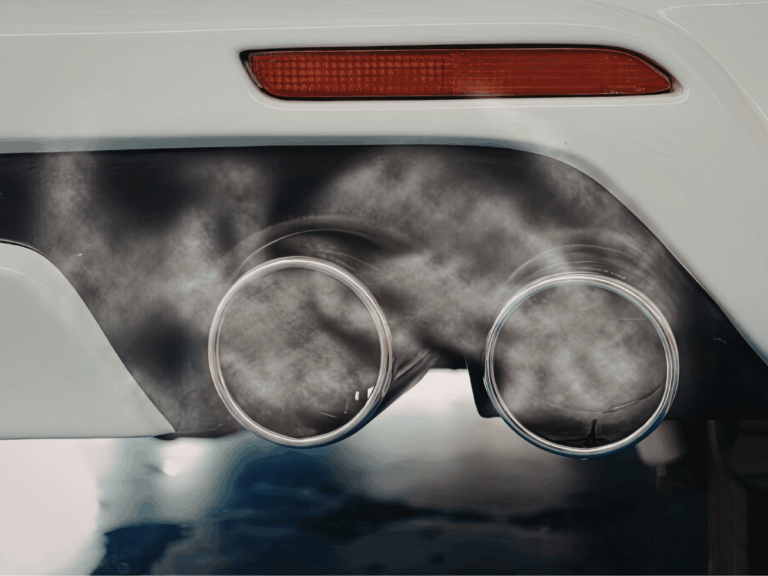 Close-up of a car’s dual exhaust pipes emitting visible fumes, highlighting the costs of engine idling, with a red tail light and part of the vehicle’s rear bumper in view.