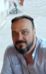A man with a beard and mustache, wearing a light blue shirt, sits outdoors on a stone-paved walkway.