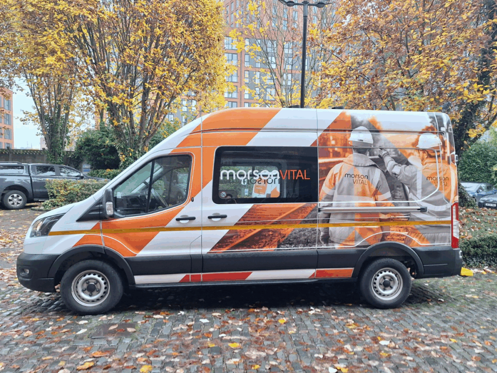 A white and orange van with "Morso Vital" branding and images of workers parked on a cobbled street, surrounded by trees with autumn foliage.