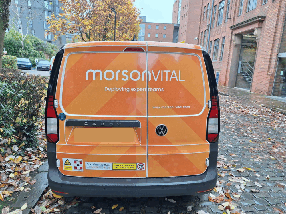 Rear view of an orange "Morson Vital" van with the slogan "Deploying expert teams" and website, parked on a cobbled street next to fallen leaves and buildings.
