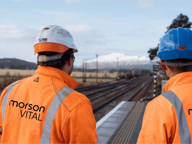Two workers in orange high-visibility jackets and helmets stand on a railway platform, looking towards railway tracks that extend into the distance.