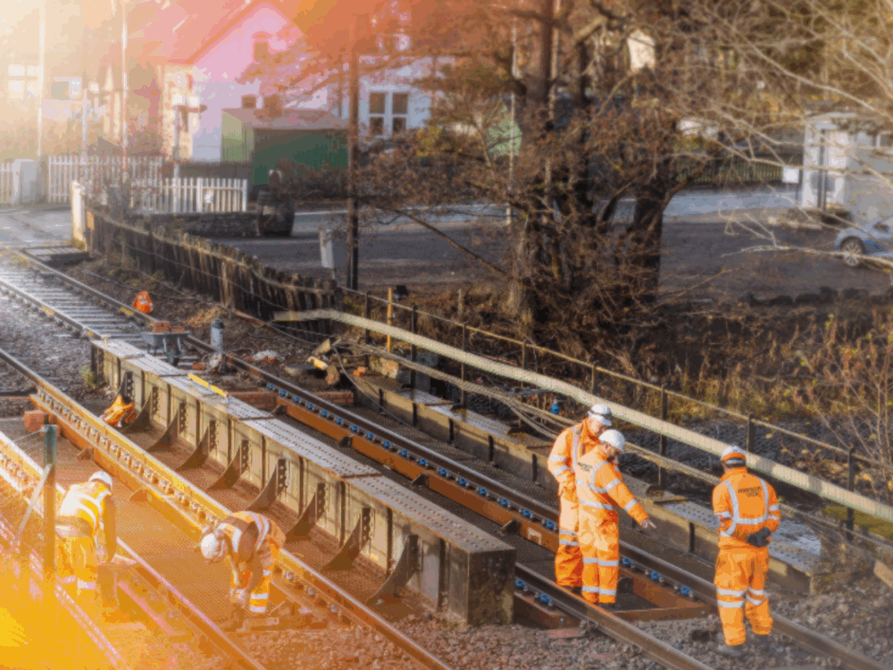 Four workers in orange safety gear inspect and repair railway tracks during daylight, with houses and trees visible in the background.