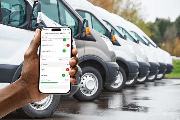 A hand holds a smartphone with a vehicle inspection checklist app displayed, in front of a row of parked white vans on a wet pavement.