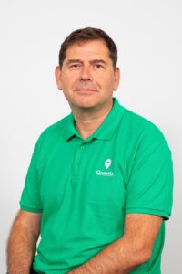 A man wearing a green polo shirt with the Quartix logo sits against a plain white background, facing the camera with a neutral expression.