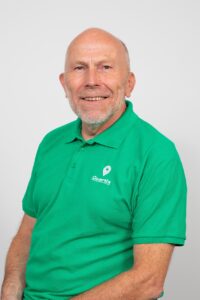 A middle-aged man with a bald head and short beard, wearing a green Quortix polo shirt, poses and smiles against a plain light background.