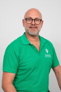 A man with glasses and a beard wearing a green polo shirt with a "Quartix" logo stands against a plain light background.