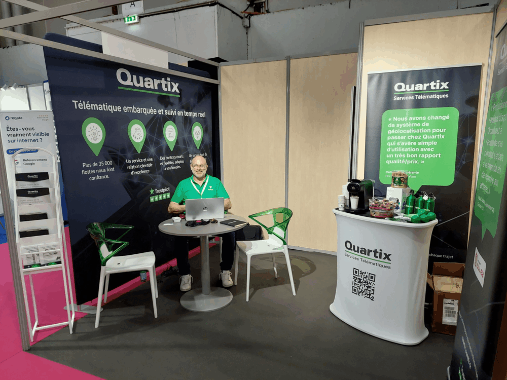 A man sits at a table in a Quartix exhibition booth, featuring banners, promotional materials, green chairs, and various branded items on display.