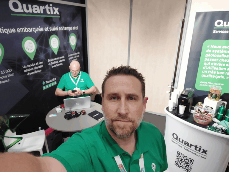 Two men in green Quartix shirts are at an exhibition booth with banners, a table, and promotional materials. One man is taking a selfie; the other is seated and looking at his phone.