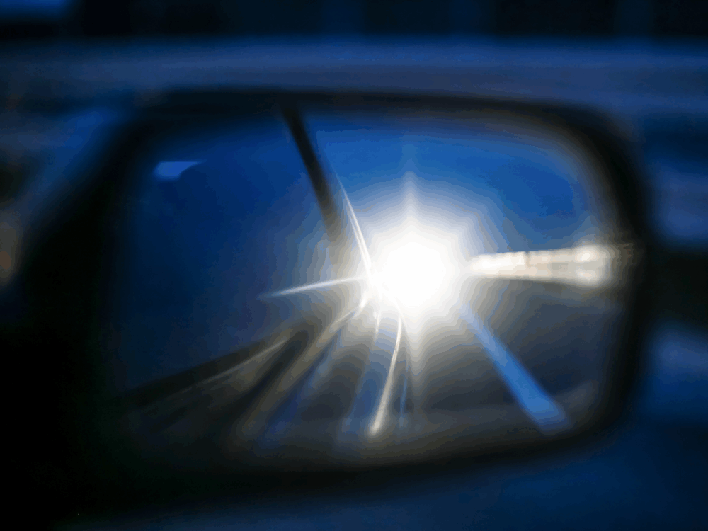 Car’s wing mirror reflecting bright headlights of a vehicle approaching from behind on a road at night.