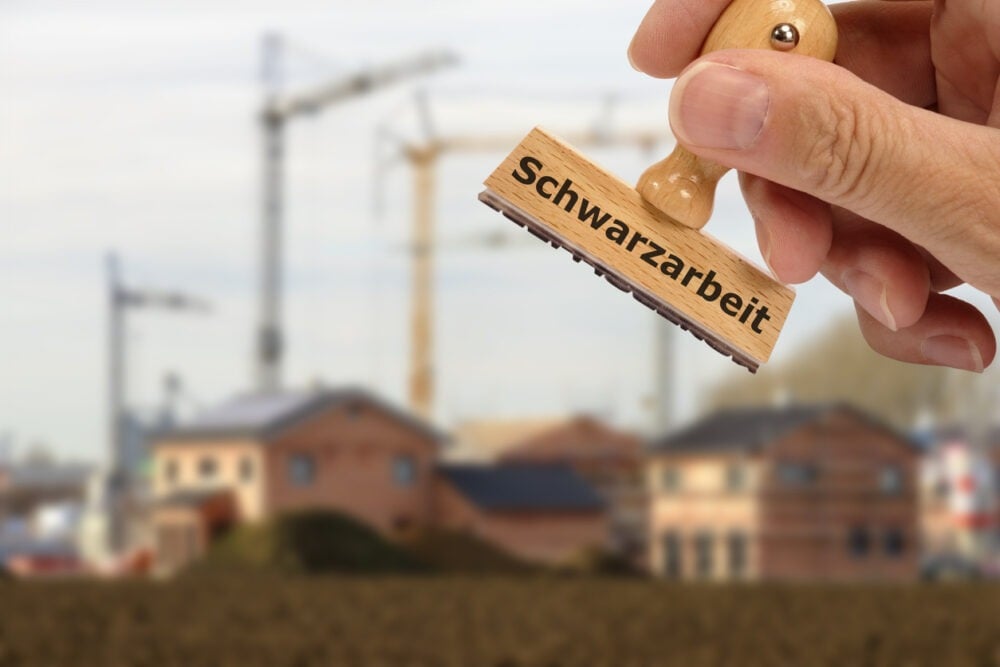 A hand holds a stamp labeled "Schwarzarbeit" with a construction site and cranes in the blurred background.