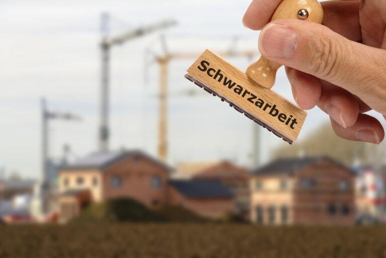 A hand holds a stamp labeled "Schwarzarbeit" with a construction site and cranes in the blurred background.
