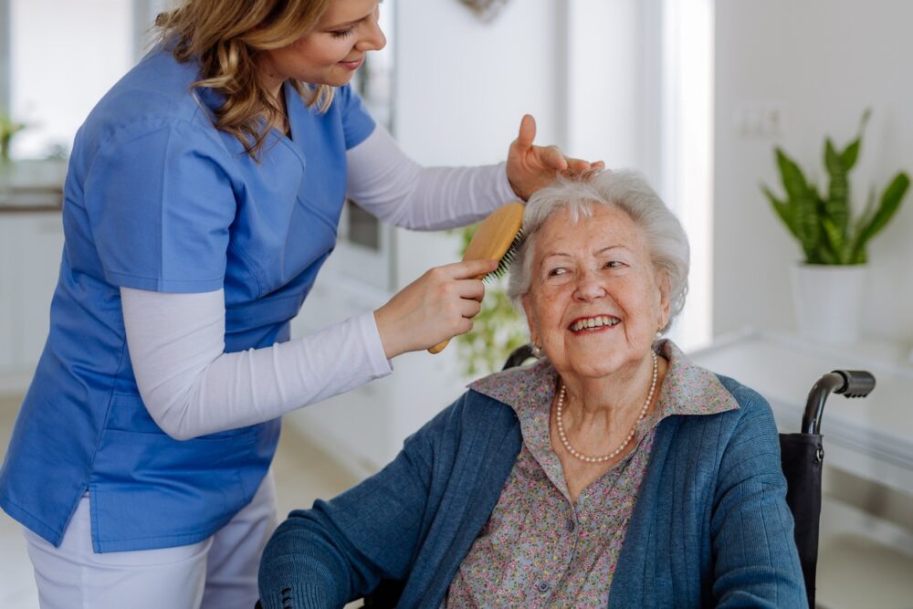 A caregiver in blue scrubs brushes the hair of an elderly woman in a wheelchair, both smiling in a bright indoor setting—demonstrating caring service that benefits from innovations like Fahrzeugortung für Pflegedienste.
