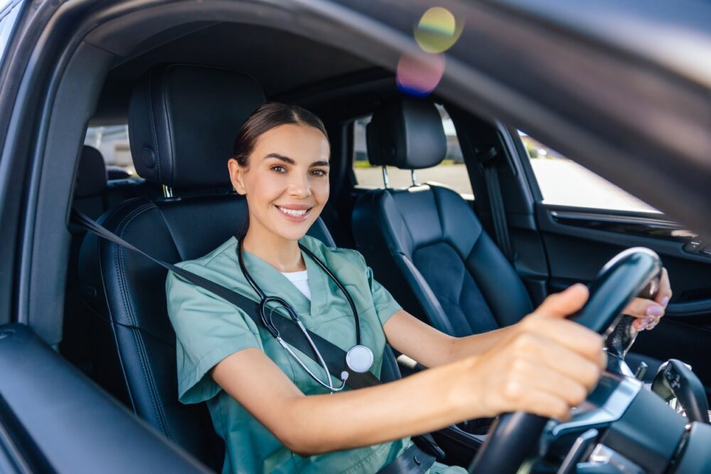 A woman wearing medical scrubs and a stethoscope sits in the driver's seat of a car, smiling at the camera—ideal for Themen wie Fahrzeugortung für Pflegedienste.