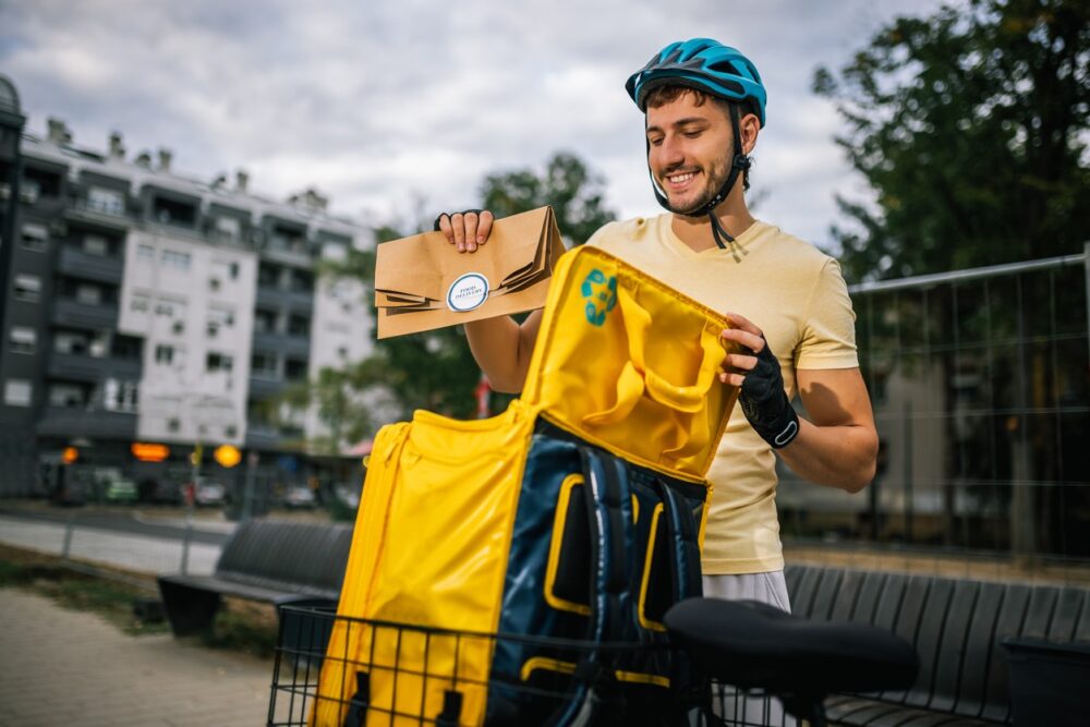 A man wearing a helmet places an envelope into a yellow delivery bag attached to a bicycle on a city street.