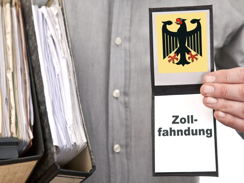 A person holds a badge displaying the German federal eagle emblem and the word "Zollfahndung" next to a file organizer with documents.