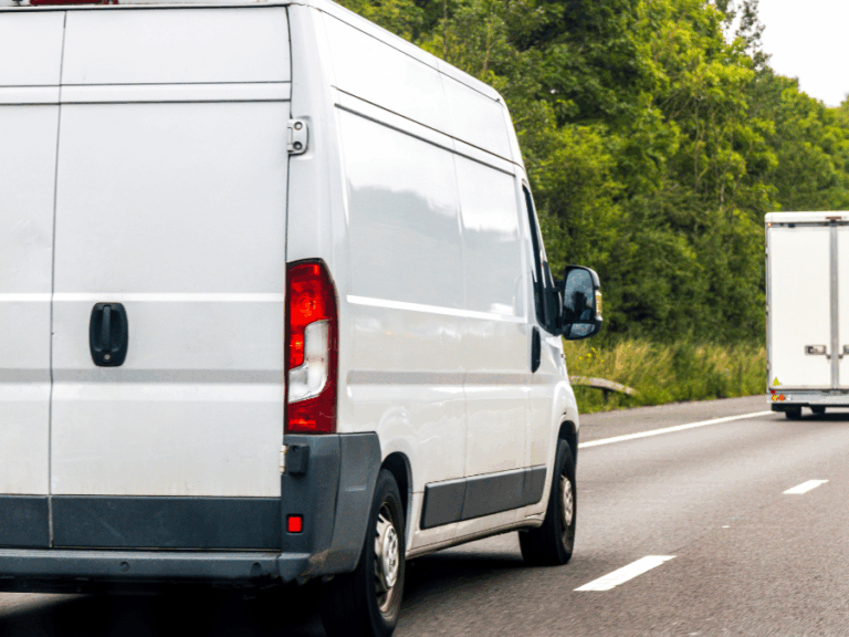 A white delivery van drives on a motorway next to a lorry, with green trees visible in the background.