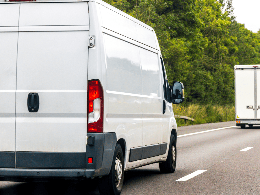 A white delivery van drives on a motorway next to a lorry, with green trees visible in the background.
