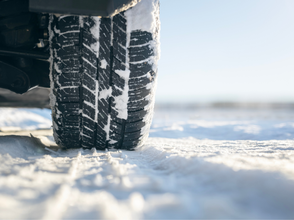 Close-up of a vehicle tyre with snow on the tread, driving on a snow-covered road in bright daylight.