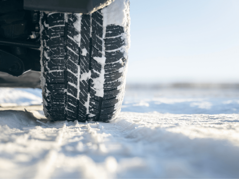 Close-up of a vehicle tyre with snow on the tread, driving on a snow-covered road in bright daylight.