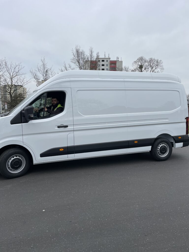 A white delivery van is parked on a paved road with a driver visible inside. Leafless trees and a multi-story building are in the background.