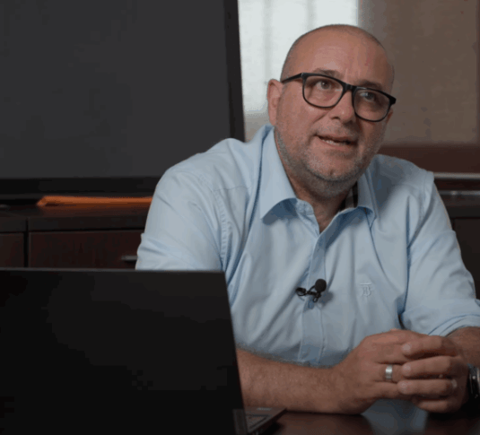 Un uomo con occhiali e barba è seduto a una scrivania con un computer portatile, indossa una camicia azzurra e parla gesticolando con le mani.