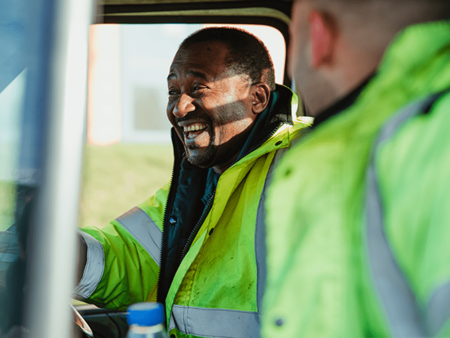 Two men wearing high-visibility jackets sit in a vehicle, with one man smiling and looking at the other.