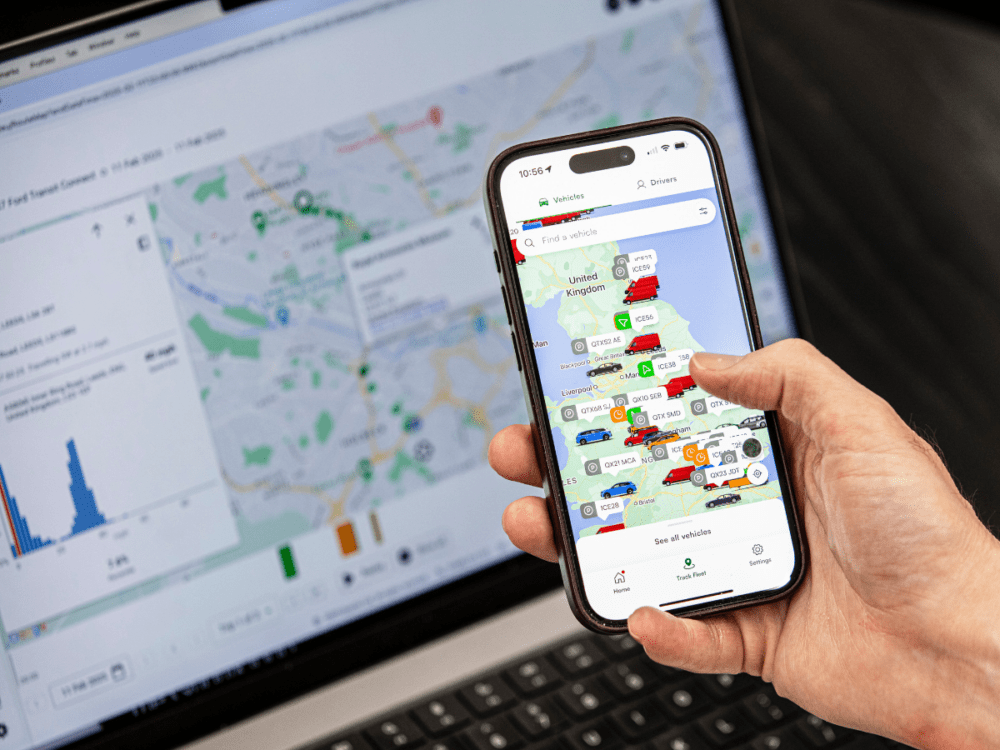 A person holds a mobile phone displaying a map with vehicle icons, while a laptop in the background shows a similar map and data charts.