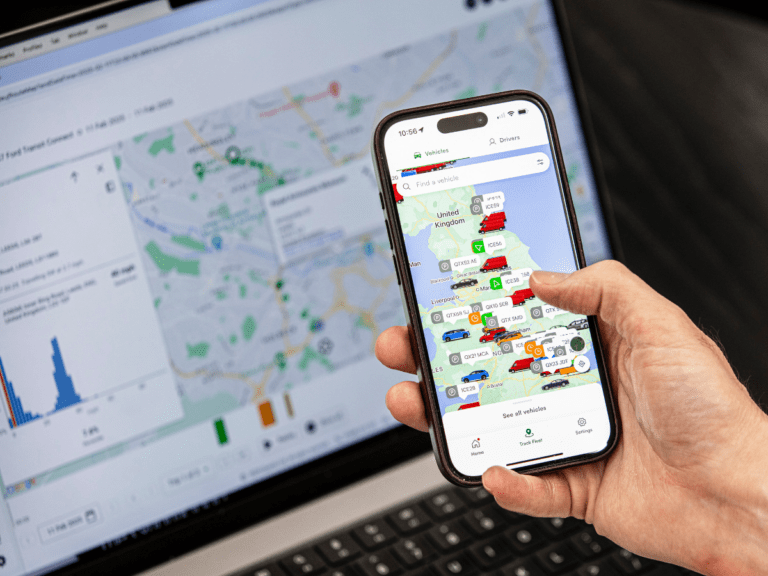 A person holds a mobile phone displaying a map with vehicle icons, while a laptop in the background shows a similar map and data charts.