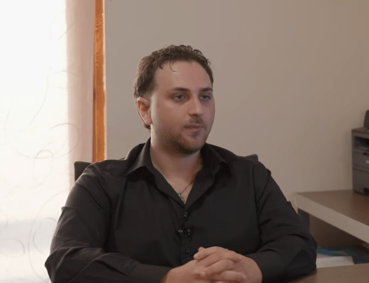 A man with short dark hair, wearing a black shirt, sits at a desk with hands clasped, looking forward. A printer and papers are visible on a side table in the background.