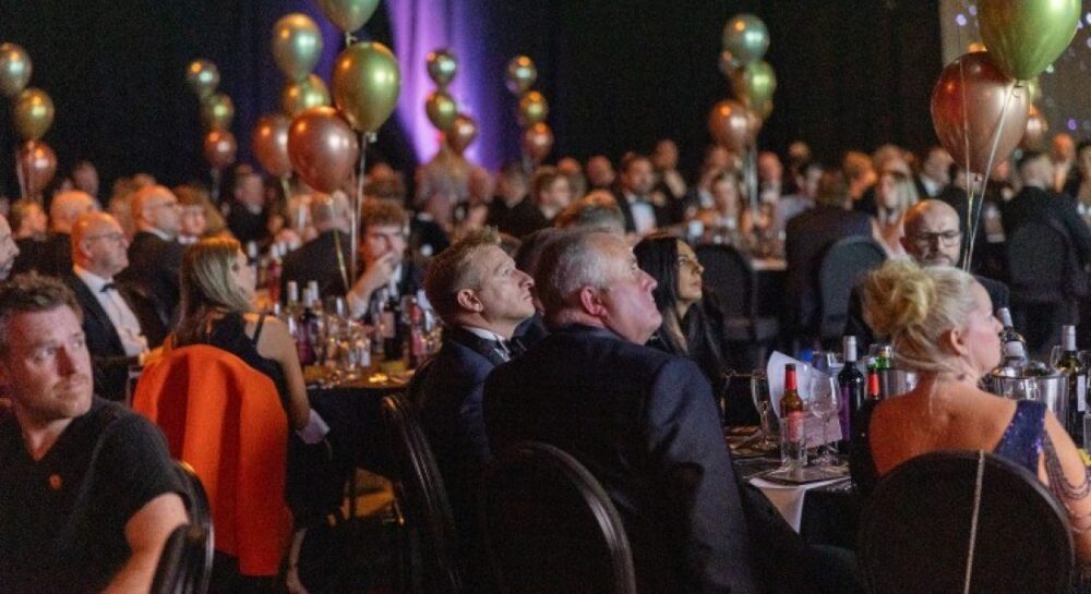 A large group of people in formal attire sit at round tables with balloons, watching an event or presentation in a dimly lit venue.