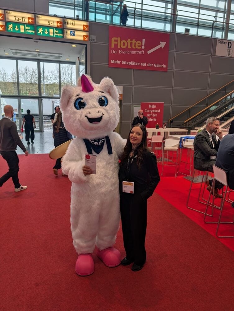 Eine Person in einem weißen Einhorn-Maskottchenkostüm steht neben einer Frau in Geschäftskleidung bei einer Indoor-Veranstaltung mit rotem Teppich und Beschilderung in deutscher Sprache.