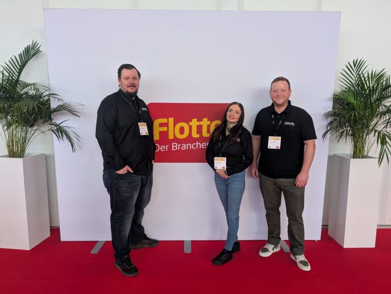 Three people stand in front of a white and red display with the text "Flotte Der Branch" at an event, all wearing black tops and name badges. Two potted plants on each side.