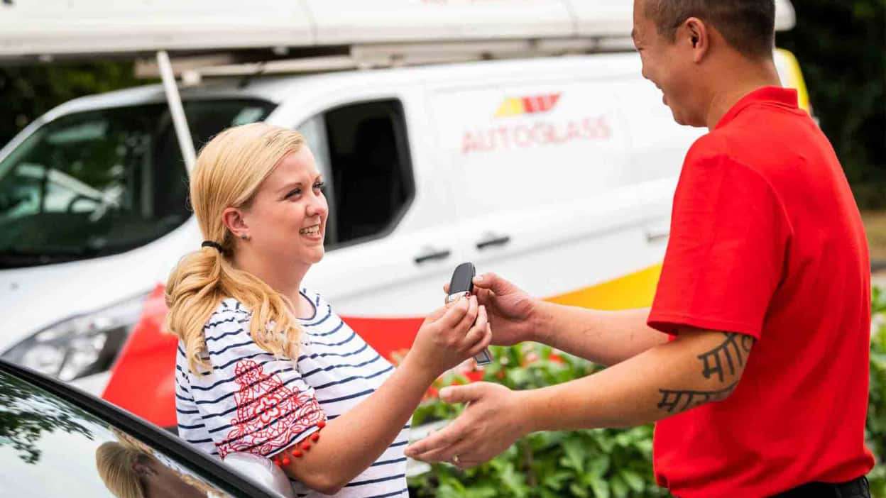 A woman receives car keys from a man in a red shirt, with an Autoglass service van in the background.